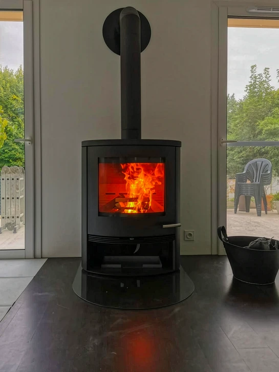 Int&eacute;rieur chaleureux avec po&ecirc;le &agrave; bois en fonctionnement, flammes visibles &agrave; travers la vitre, r&eacute;serve de b&ucirc;ches int&eacute;gr&eacute;e dans la partie basse