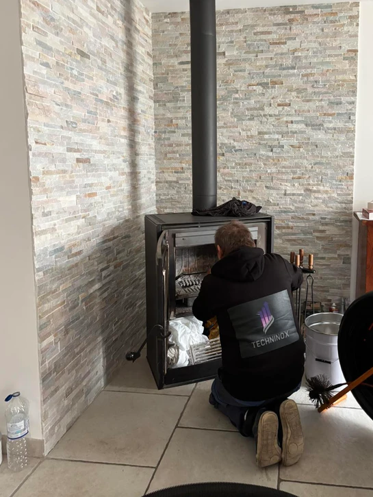 Photographie professionnelle montrant un artisan en tenue de travail inspectant ou travaillant sur plusieurs types d'équipements de chauffage distincts dans un espace de travail ou d'intervention : poêle à bois, chaudière gaz, conduit de fumée, et granulés. L'image doit transmettre l'expertise technique, la polyvalence et la maîtrise professionnelle des différents systèmes de chauffage, sans ambiance résidentielle ni mise en scène décorée.